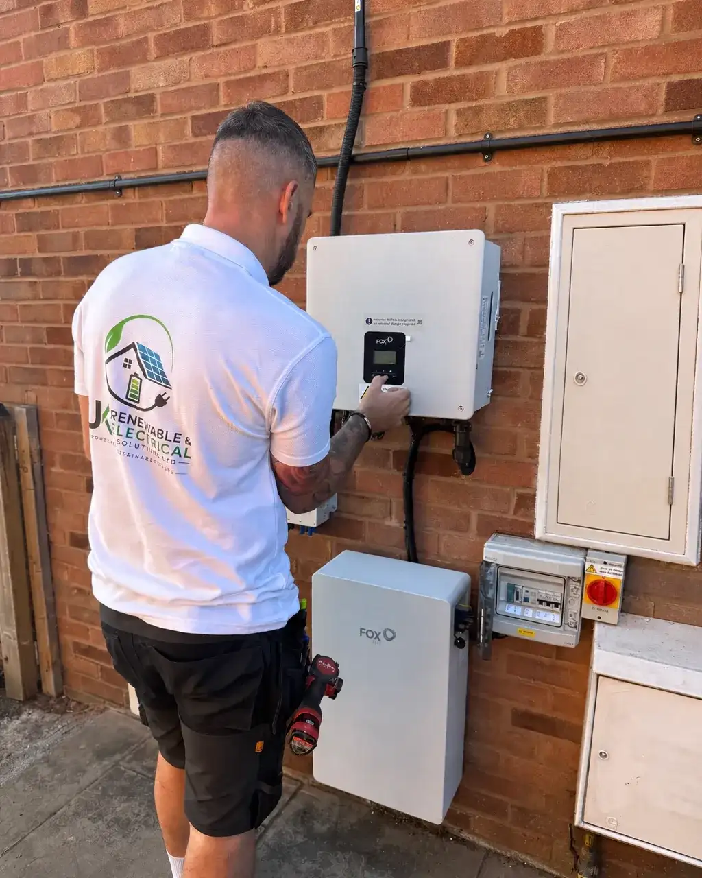 Electrician in a white company polo shirt checking a white gloss meter cupboard beside a battery system